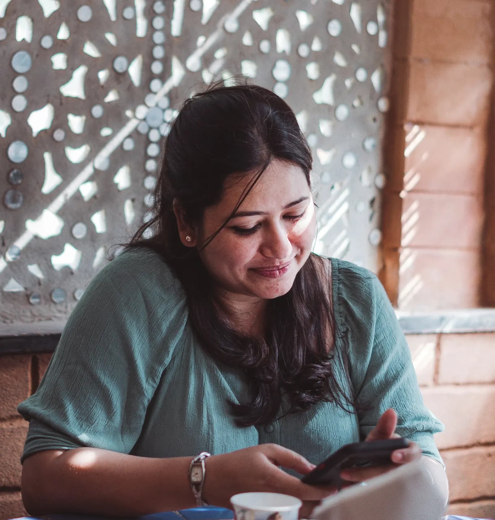 A woman in a green top smiles at her phone at a table, a cut-stone screen and brick wall behind her.
